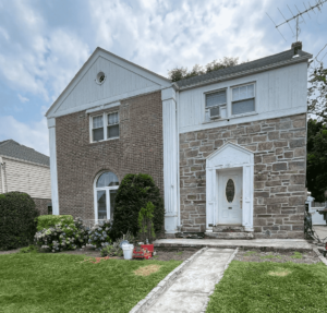 A two-story house with a white and brick facade, stone entryway, and overgrown yard.