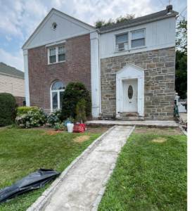 A two-story house with a white and brick facade, stone entryway, and overgrown yard.