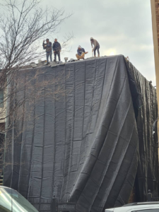 Four workers on the roof of a building covered with a large black tarp.